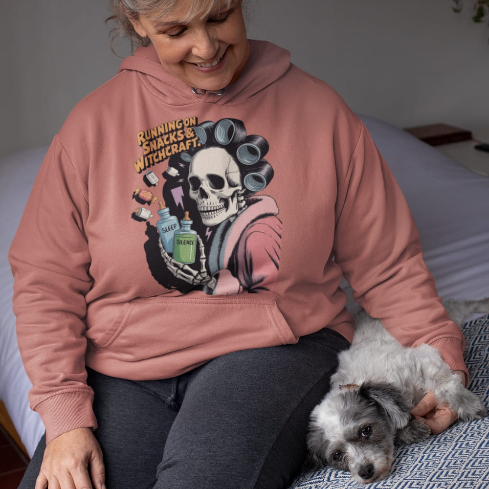 Person wearing a pink hoodie with a skeleton design, sitting on a bed with a small dog.