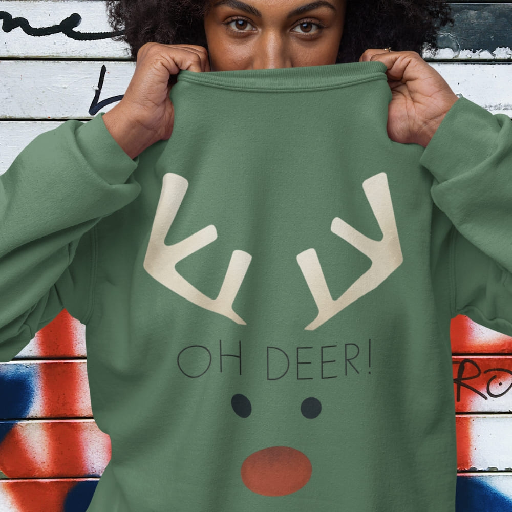 Person holding a green sweatshirt with reindeer design and 'OH DEER!' text against a graffiti-covered wall.