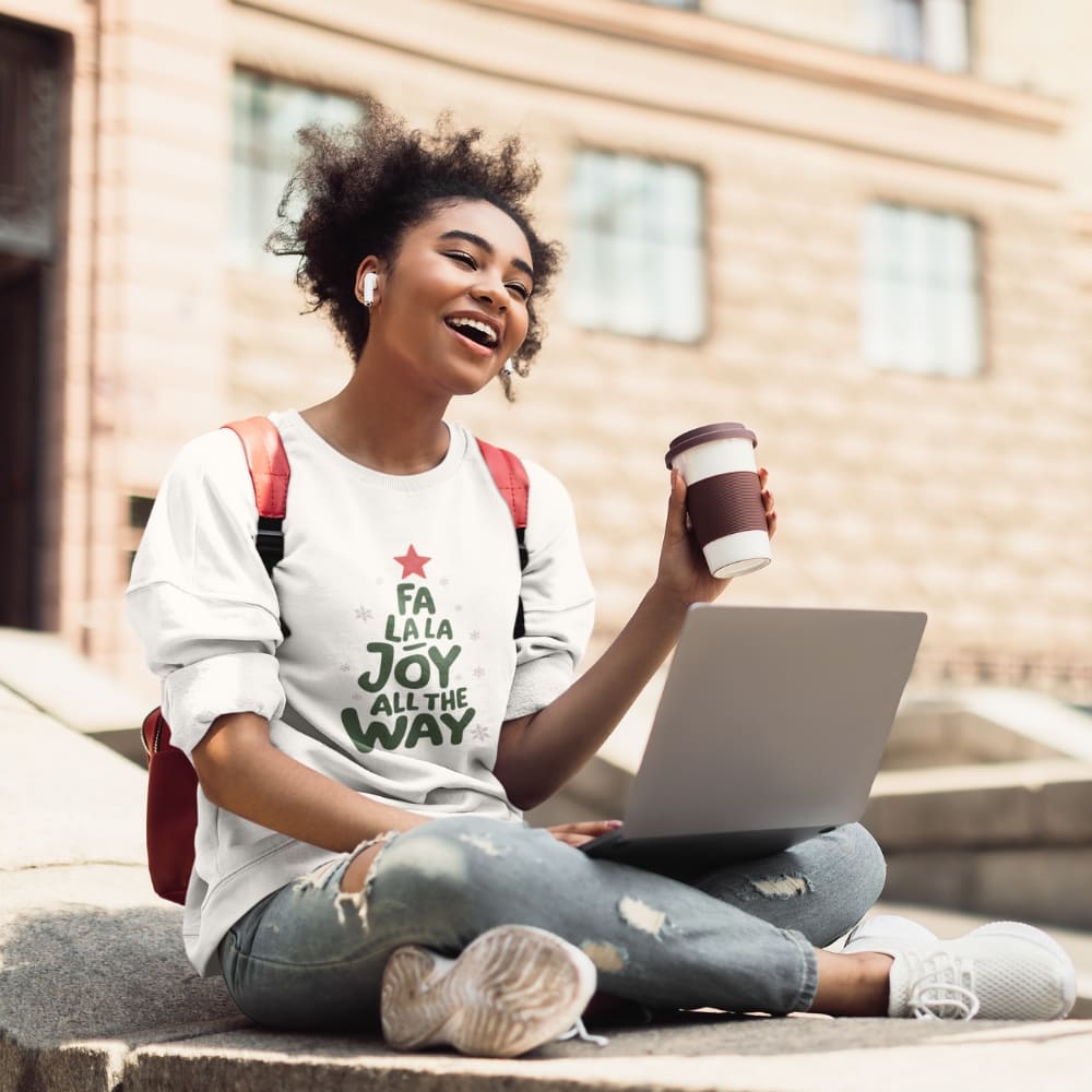 Woman sitting outdoors with a laptop and coffee, wearing a shirt with text.