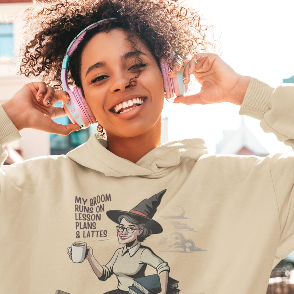 Woman wearing a hoodie with a graphic design of a witch holding a coffee cup, with text 'My broom runs on lesson plans & lattes'.