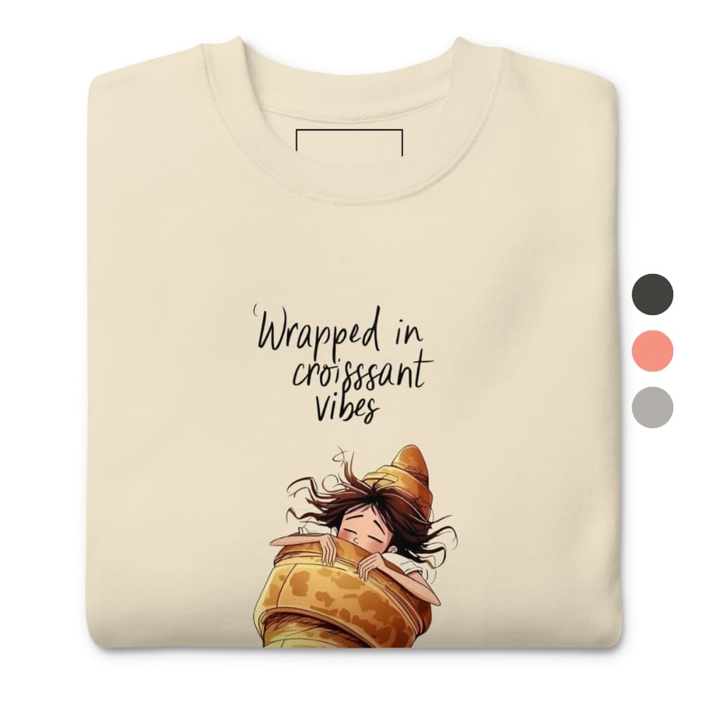Beige t-shirt with cartoon character wrapped in a croissant and text 'Wrapped in croissant vibes' on a white background.