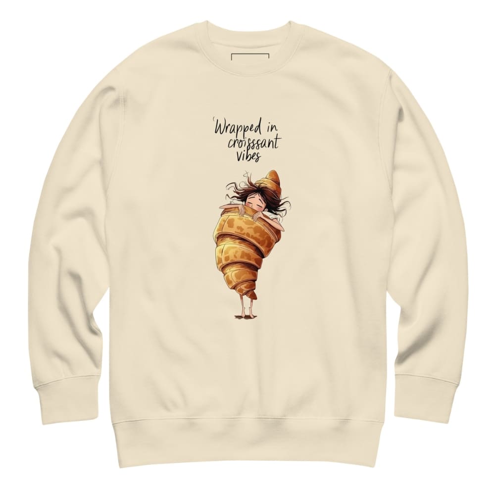 Beige sweatshirt with a cartoon character wrapped in a croissant and text 'Wrapped in croissant vibes' on a white background