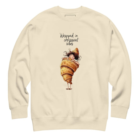 Beige sweatshirt with a cartoon character wrapped in a croissant and text 'Wrapped in croissant vibes' on a white background