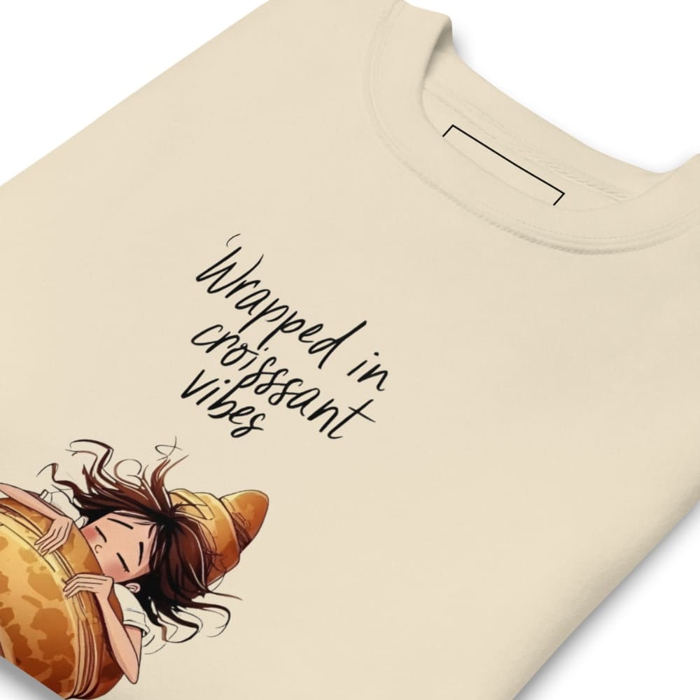 Beige t-shirt with an illustration of a person wrapped in a croissant and text 'Wrapped in croissant vibes' on a white background.