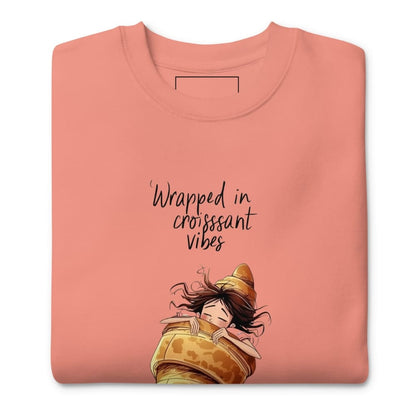 Folded pink sweatshirt with cartoon character and text 'Wrapped in croissant vibes' on a white background