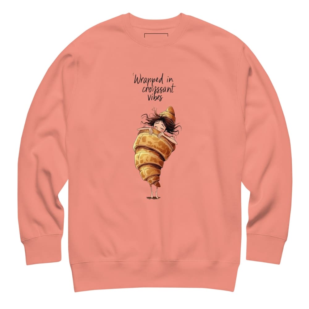 Coral sweatshirt with a cartoon character wrapped in a croissant and text 'Wrapped in croissant vibes' on a white background.