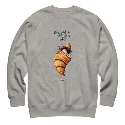 Gray sweatshirt with a cartoon character wrapped in a croissant and text 'Wrapped in croissant vibes' on a white background