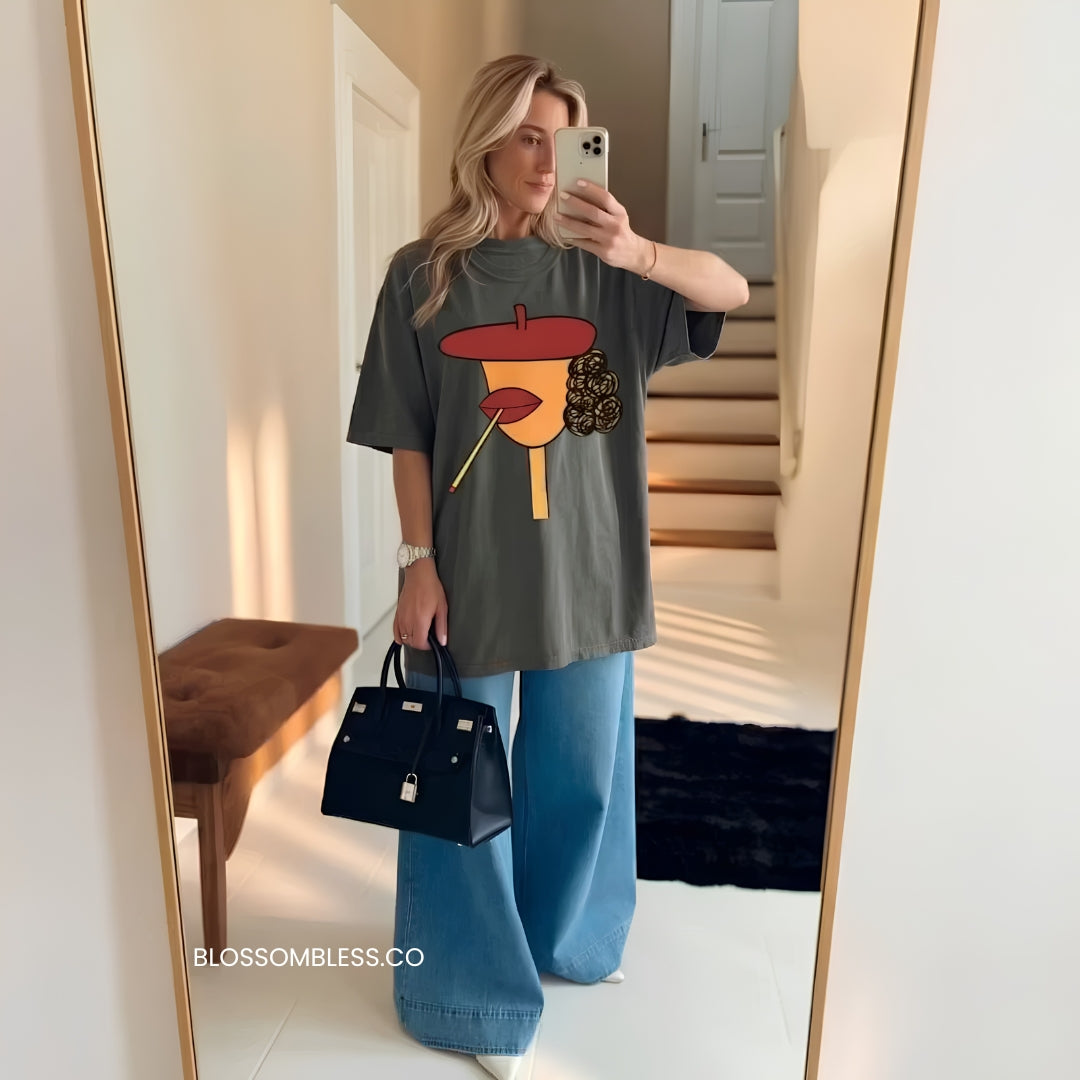 A woman takes a mirror selfie in a home setting, wearing an oversized dark gray graphic t-shirt with a colorful pop-art design, wide-leg blue jeans, and carrying a black handbag.