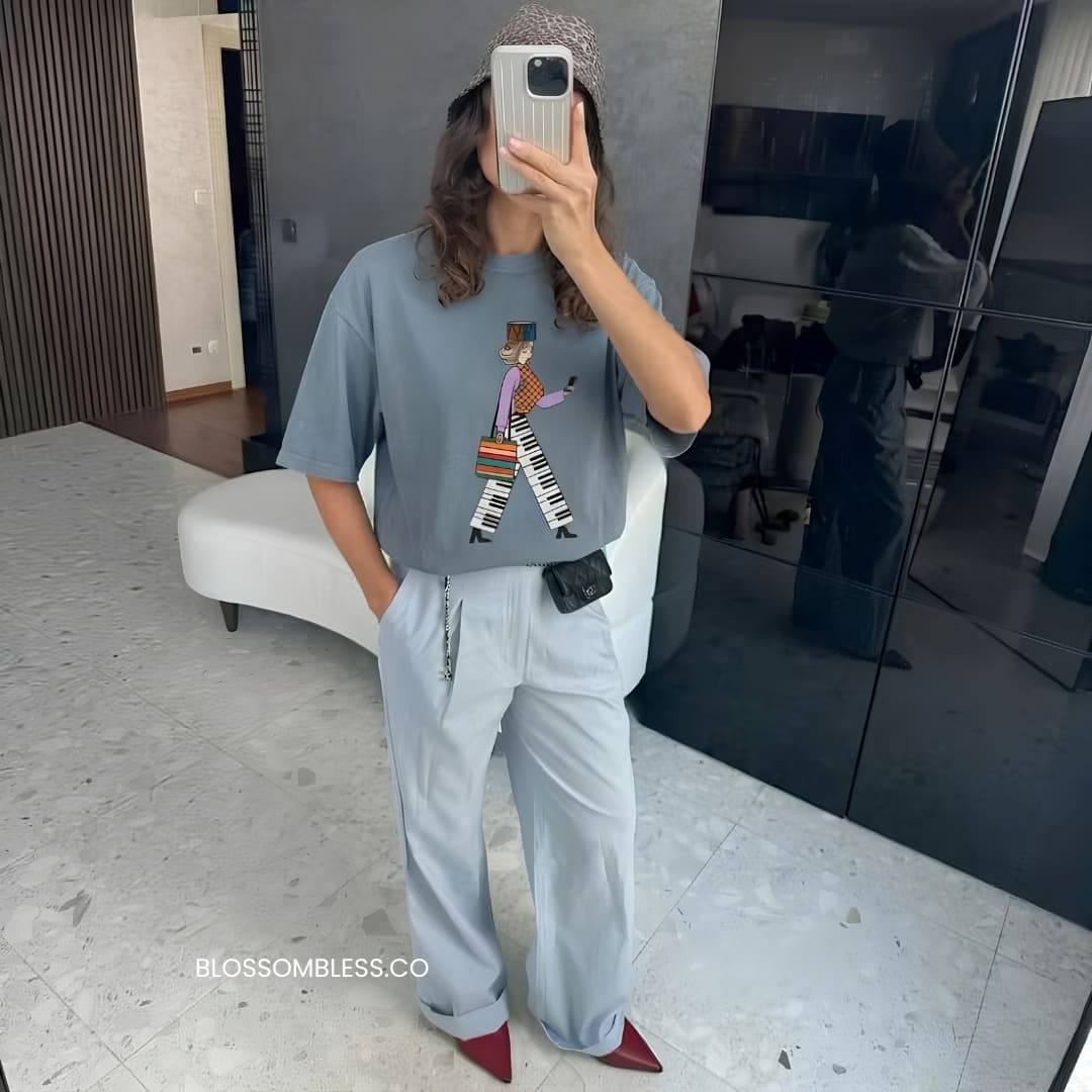 A person taking a mirror selfie in a modern room, wearing a light blue-gray graphic t-shirt with a cartoon character on the front, matching wide-leg trousers, and red pointed shoes. The watermark 'BLOSSOMBLESSE.
