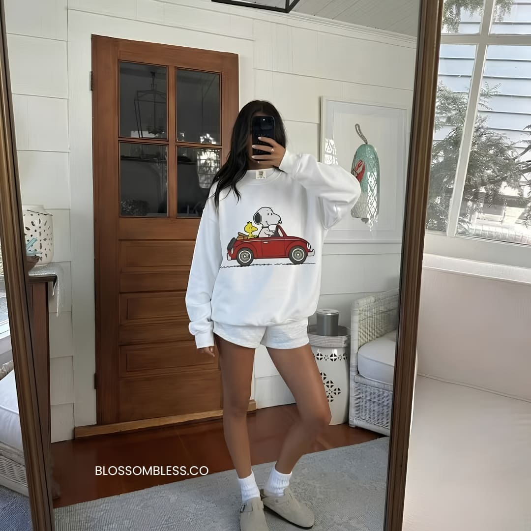 A woman takes a mirror selfie wearing a white crewneck sweatshirt with a graphic of Snoopy in a red car, paired with white shorts, white socks, and beige sneakers. The background shows a home interior with a wooden door and white paneled walls.