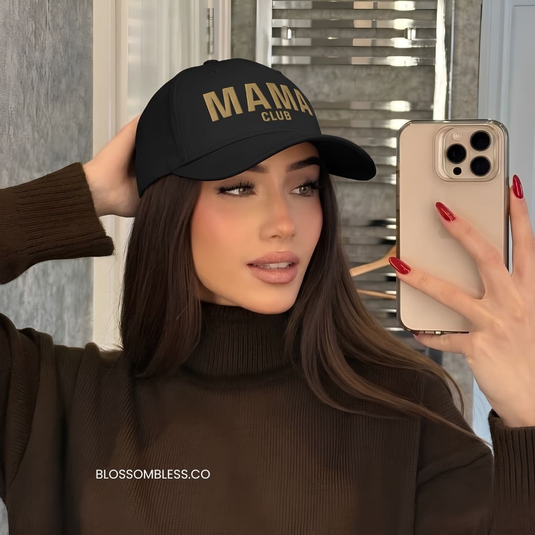 A woman taking a mirror selfie wearing a black baseball cap with gold embroidered text that reads 'MAMA CLUB'. She is wearing a dark brown ribbed turtleneck sweater and holding a beige smartphone.