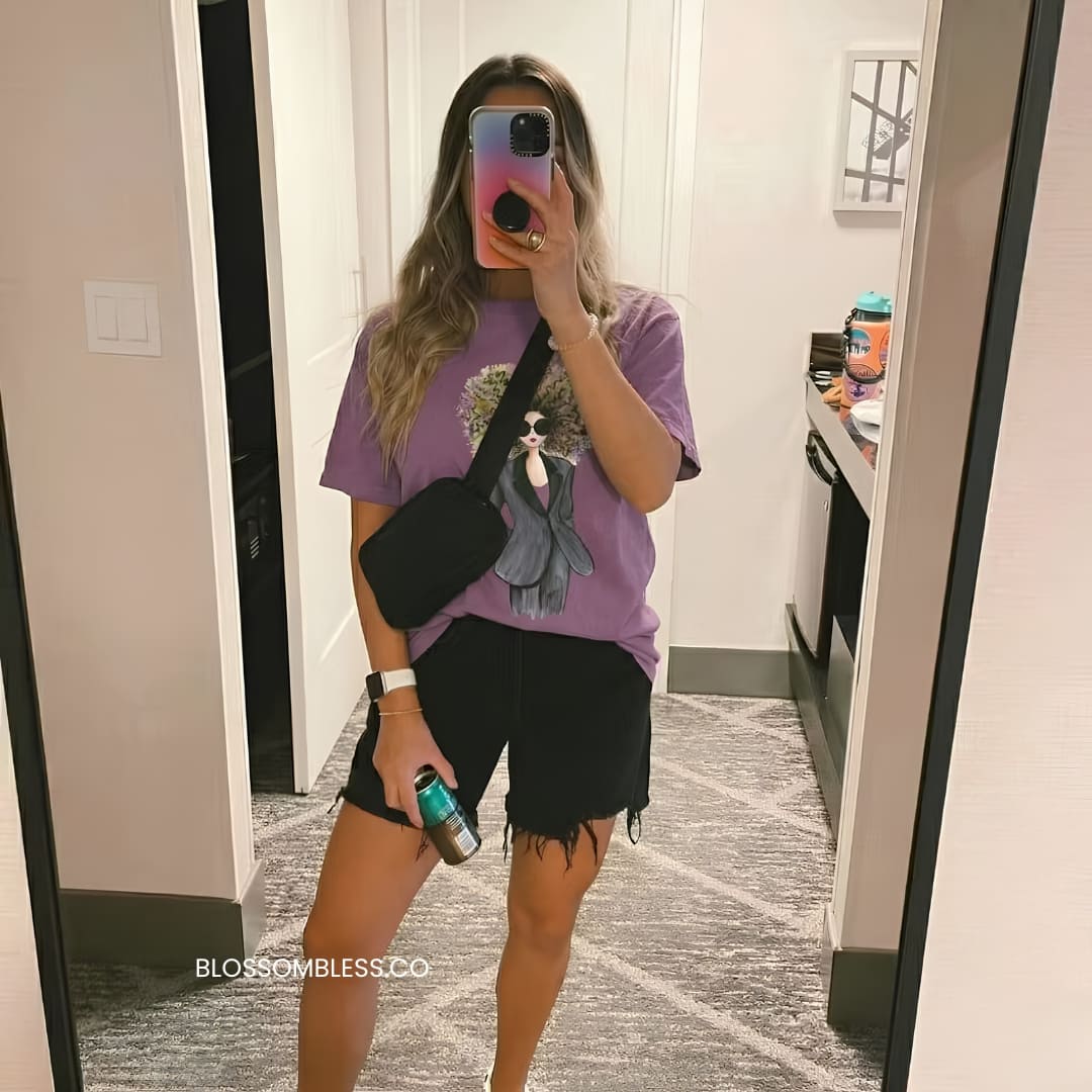 A woman takes a mirror selfie in a hotel or apartment hallway wearing a purple graphic t-shirt, black denim shorts, and a black crossbody bag. The t-shirt features a graphic of a woman in a gray dress with a pink and blue gradient phone case.