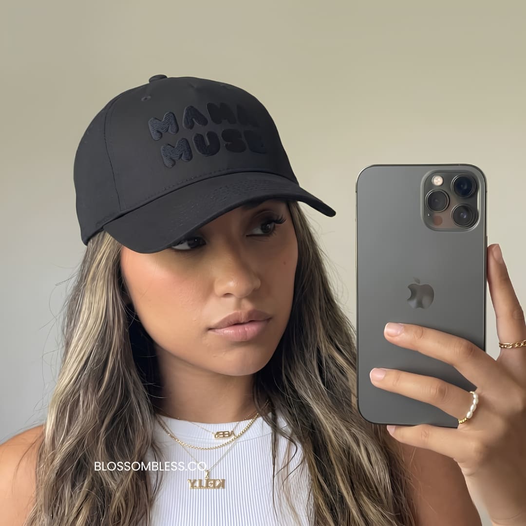 A woman takes a mirror selfie wearing a black baseball cap with 'MADAME MUSE' embroidered in tonal black across the front. She is holding a dark gray iPhone next to her face.