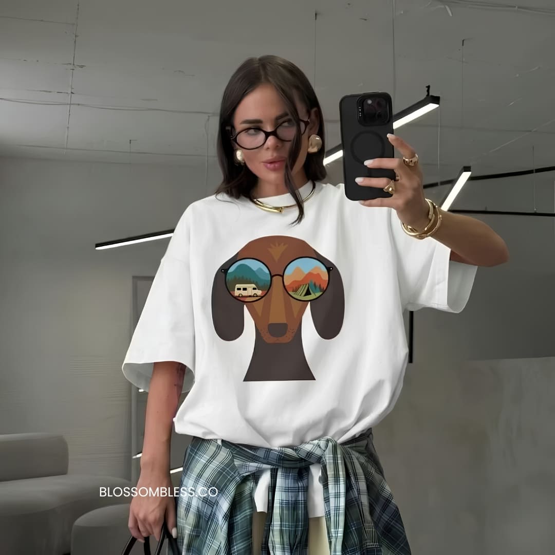 A woman takes a mirror selfie wearing a white oversized t-shirt with a graphic of a brown dog wearing sunglasses with landscape reflections, paired with a green plaid shirt tied at the waist. The brand name 'BLOSSOMBLES.