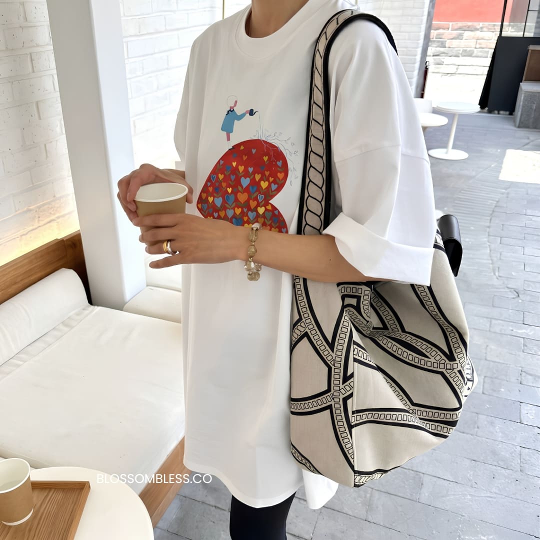 A person wearing a white oversized graphic T-shirt and carrying a large cream tote bag with black geometric patterns, standing in a bright indoor cafe-like setting.