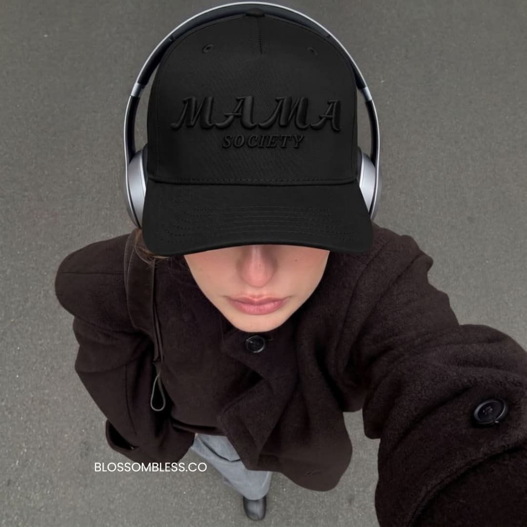 A person wearing a black baseball cap with embossed 'MAMA SOCIETY' text, silver over-ear headphones, and a dark brown coat, photographed from above on a gray paved surface.