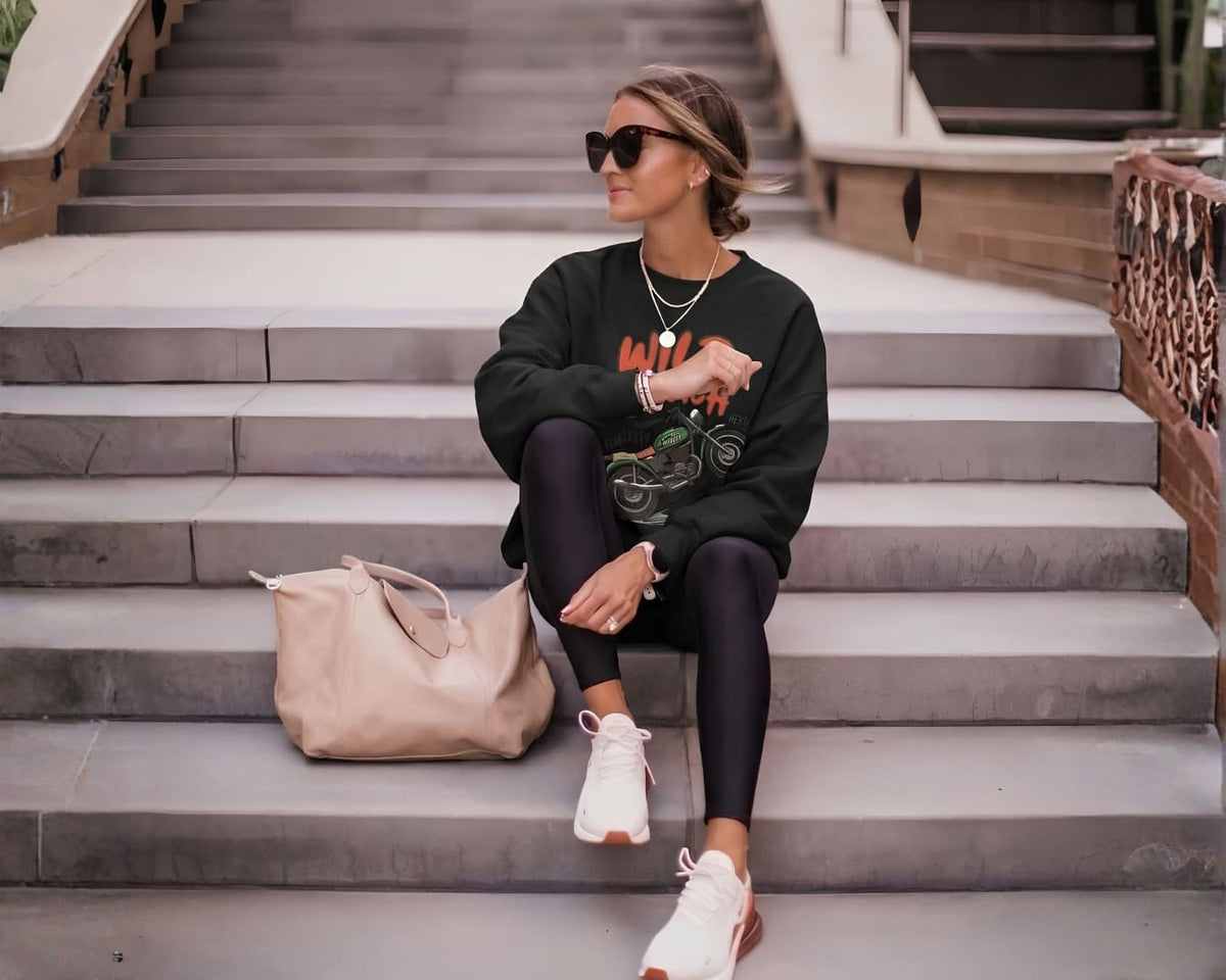 Woman in sunglasses and a black graphic sweatshirt sitting on outdoor stone steps beside a blush tote bag