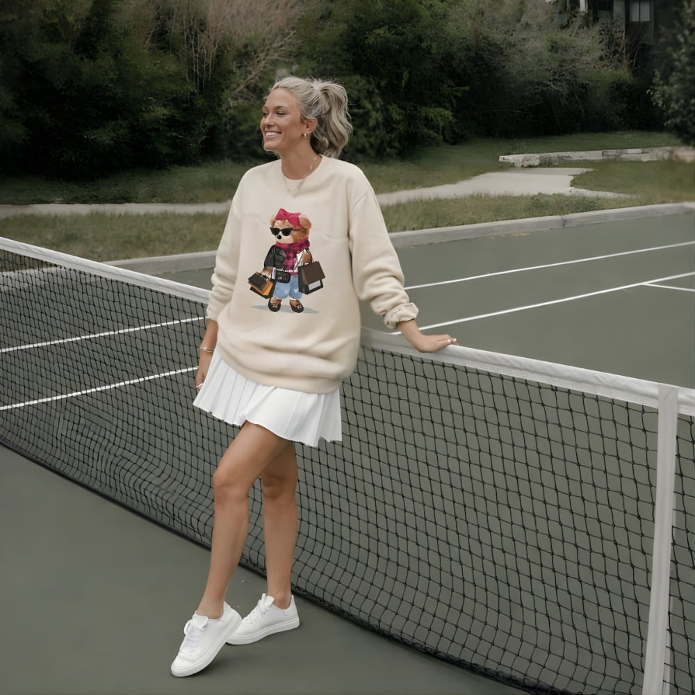 Woman in a beige sweatshirt with a graphic design, white skirt, and white sneakers standing on a tennis court.