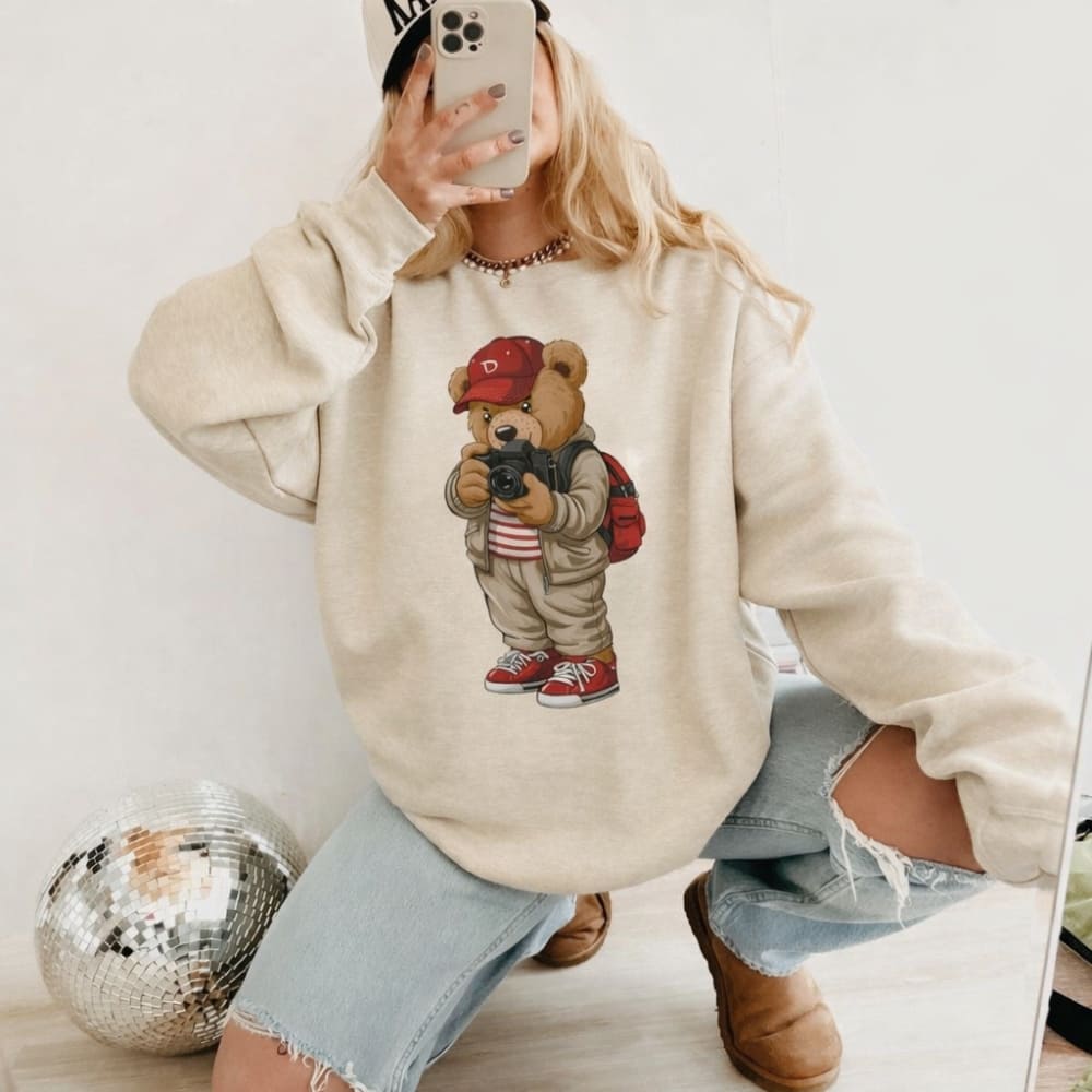 Person wearing a beige hoodie with a teddy bear graphic, taking a mirror selfie.