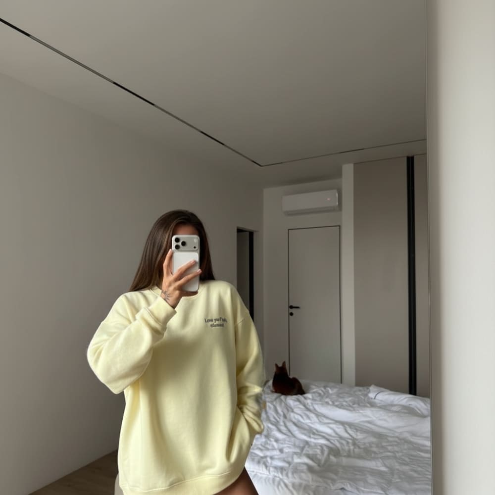 Person wearing a light yellow sweatshirt taking a mirror selfie in a room with a bed and a cat.