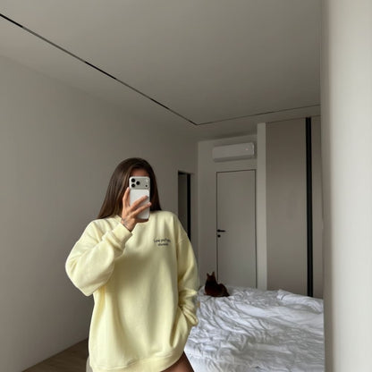 Person wearing a light yellow sweatshirt taking a mirror selfie in a room with a bed and a cat.