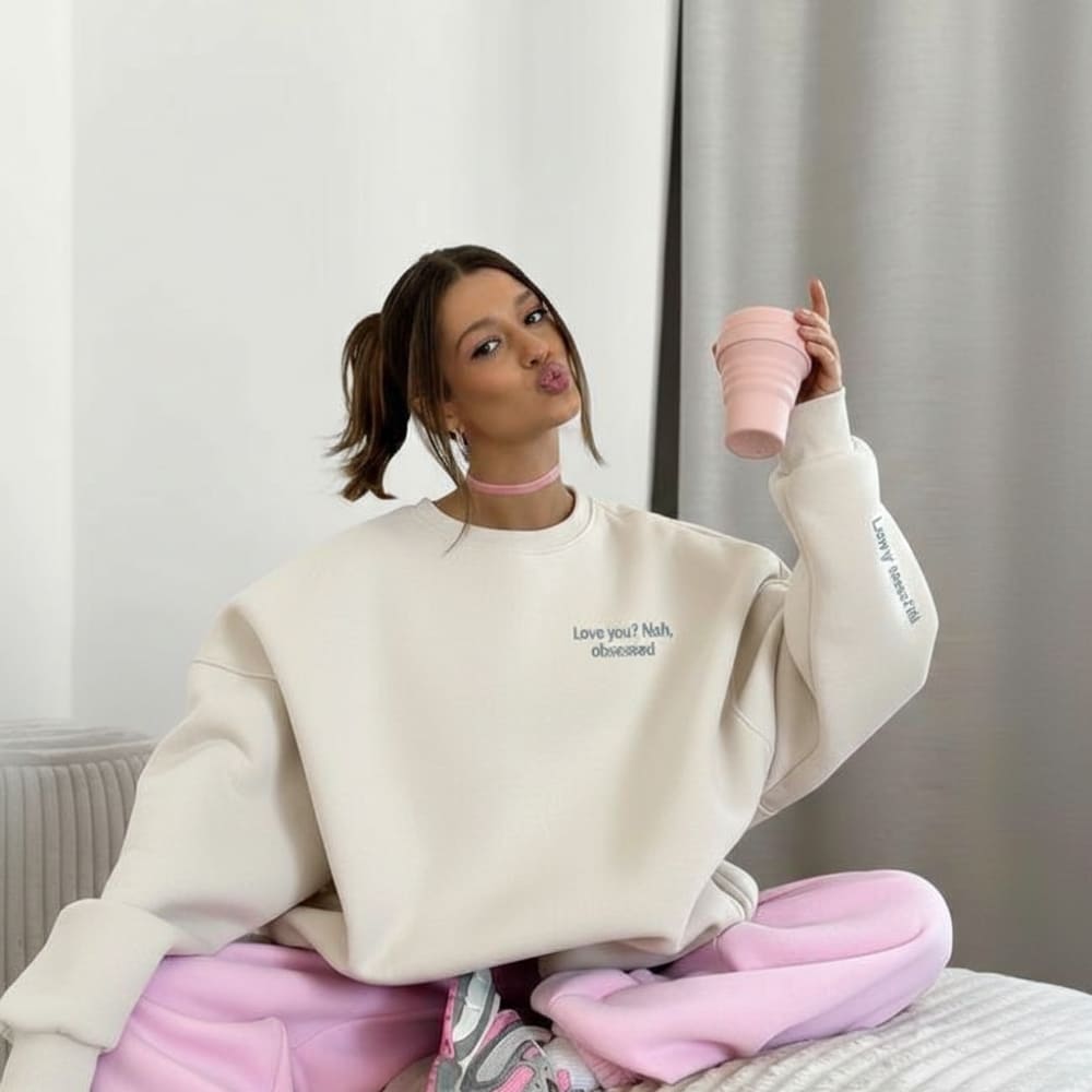 Woman wearing a beige sweatshirt and pink pants, holding a pink mug indoors.