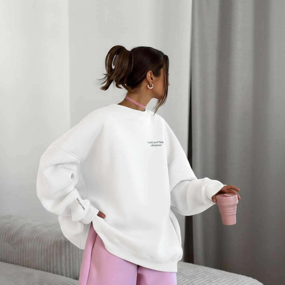 Person wearing a white sweatshirt with text, holding a pink cup in a minimalistic room.