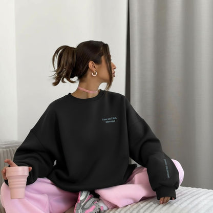 Person wearing a black sweatshirt with text, sitting on a bed holding a pink cup.