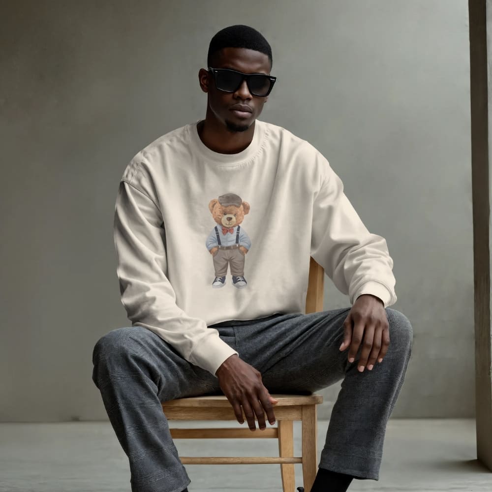 Man wearing a white sweatshirt with a teddy bear graphic, sitting on a wooden stool against a gray background.