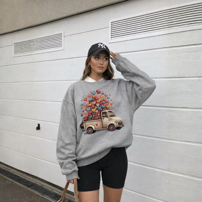 Person wearing a gray sweatshirt with a colorful truck and heart design, standing against a white wall.