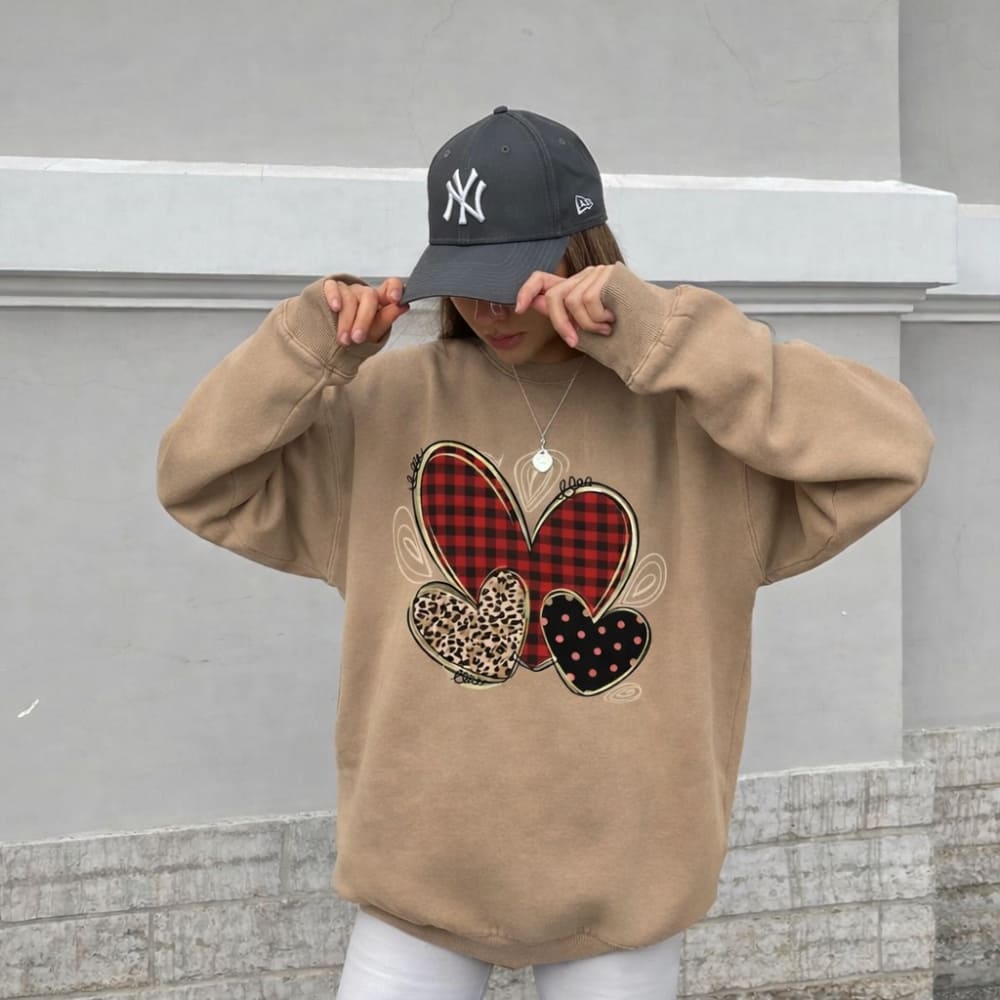 Person wearing a beige sweatshirt with heart designs and a black cap in front of a gray wall.