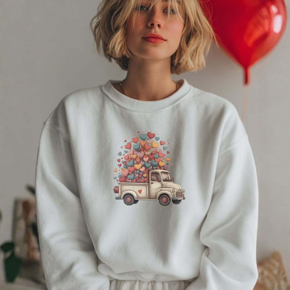 Person wearing a white sweatshirt with a colorful truck and heart design, standing indoors.