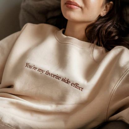 Person wearing a beige sweatshirt with text 'You're my favorite side effect' in a casual setting.