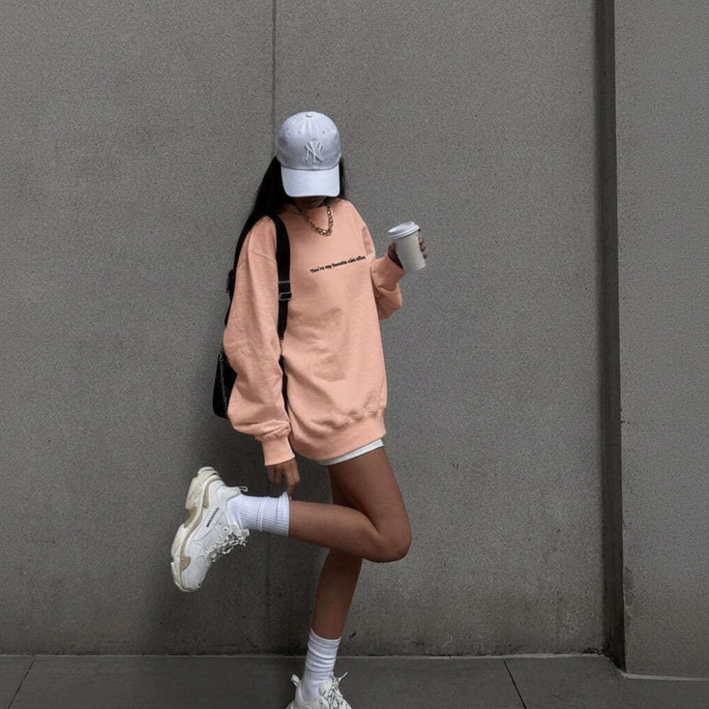 Person in a pink sweatshirt and white sneakers standing against a gray wall.