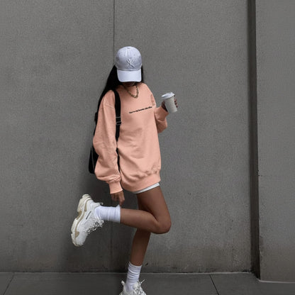 Person in a pink sweatshirt and white sneakers standing against a gray wall.