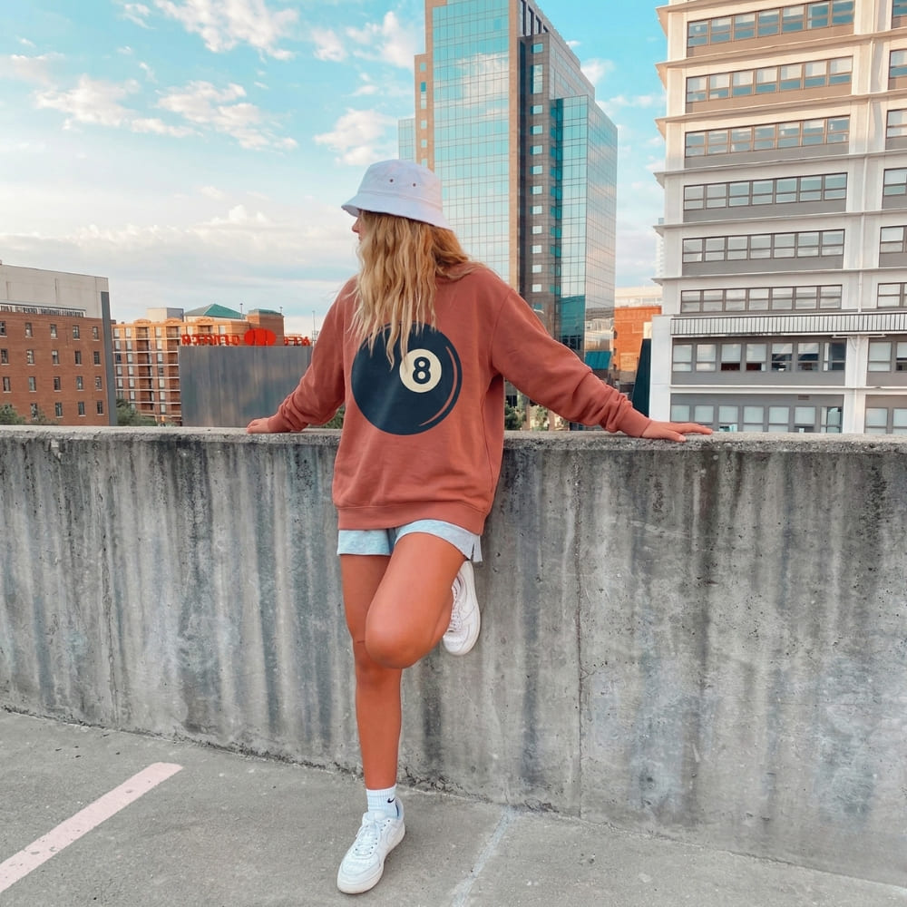 Person wearing a red sweatshirt with a blue circle design, white sneakers, and a light blue cap on a rooftop with cityscape background.