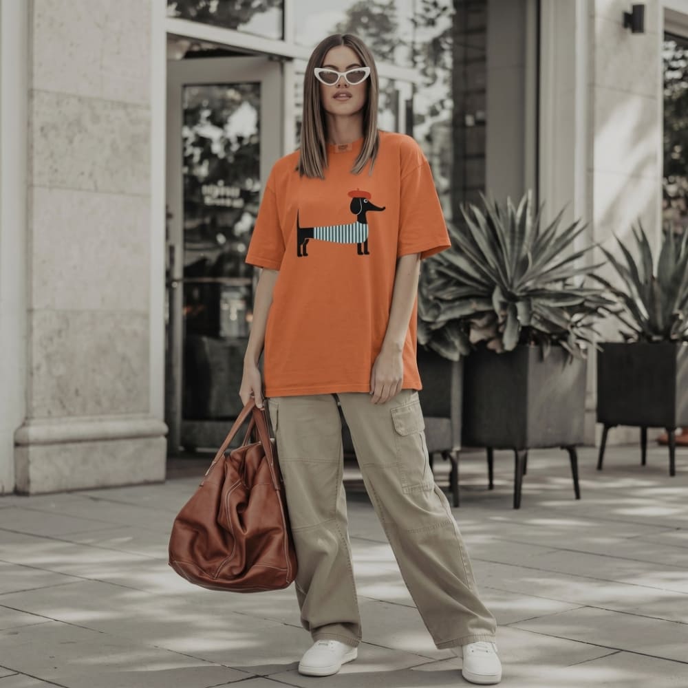 Person wearing an orange t-shirt with a dog graphic, holding a brown bag, standing on a sidewalk.