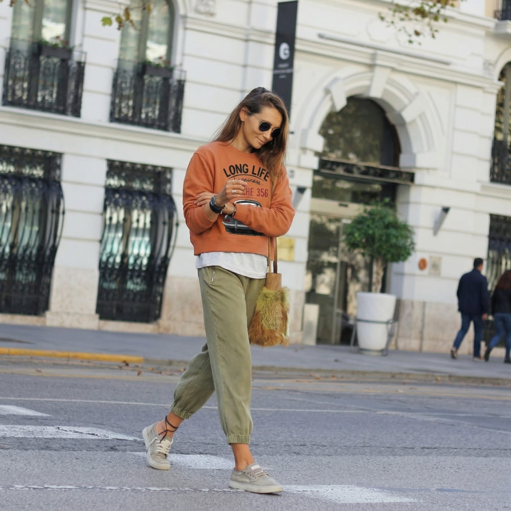 Woman walking on a street wearing an orange sweatshirt and green pants.