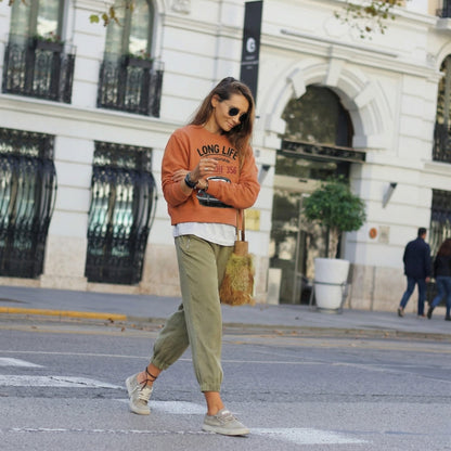 Woman walking on a street wearing an orange sweatshirt and green pants.