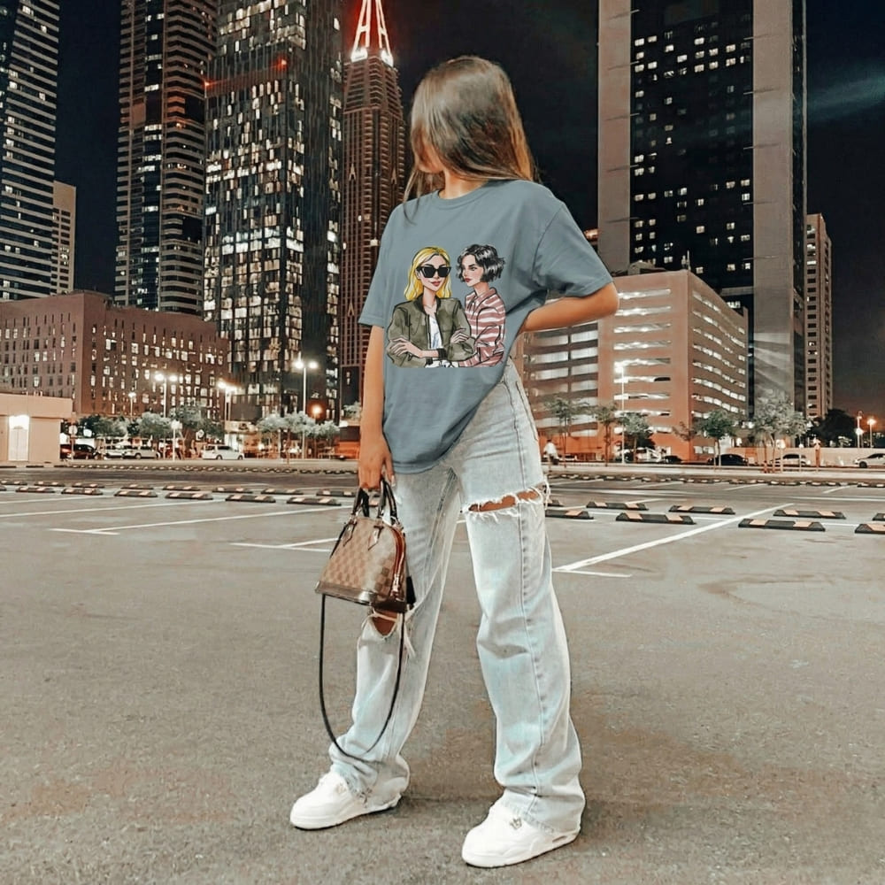 Person wearing a graphic ice-blue t-shirt and white jeans standing on a city street at night.