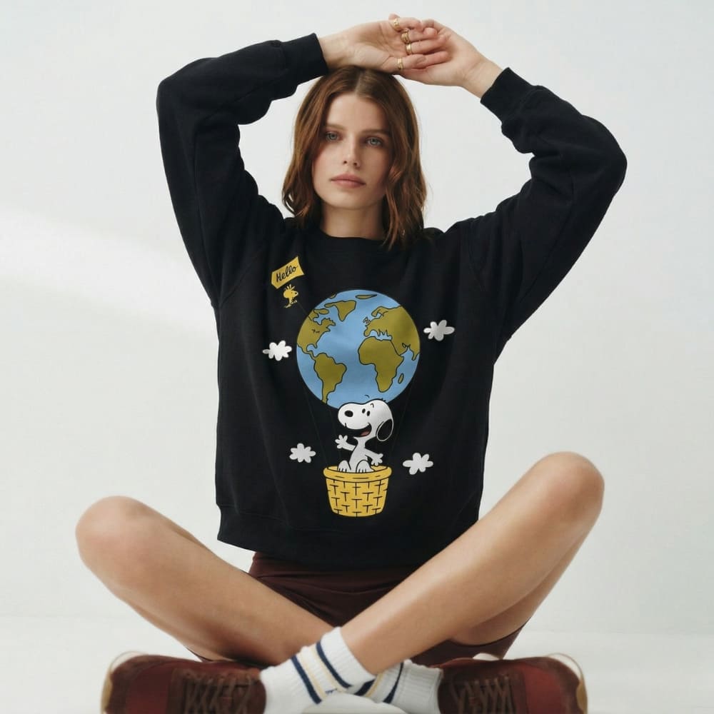 Person wearing a black sweatshirt with a cartoon design of Snoopy and a globe, sitting on a white background.