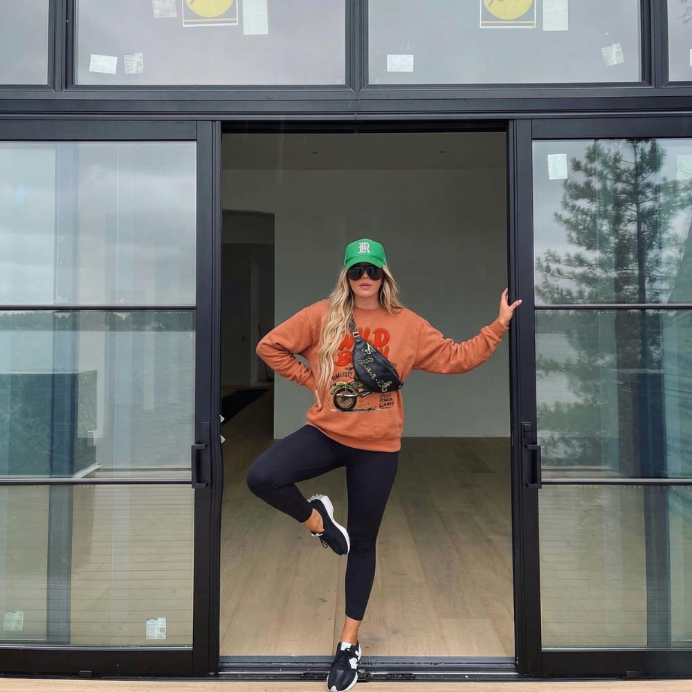 Person posing in front of a glass door wearing an orange sweatshirt and black leggings.