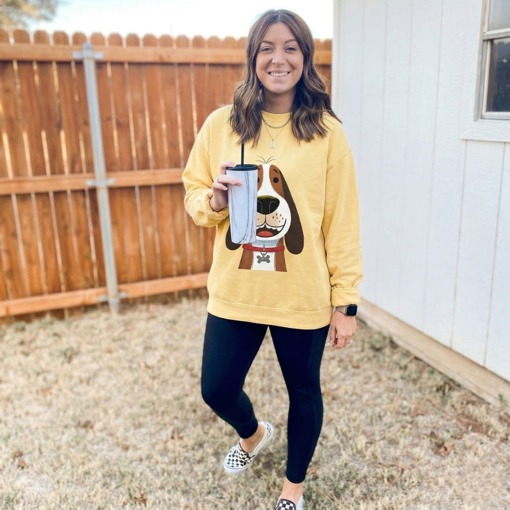 Person wearing a yellow sweatshirt with a dog graphic, holding a drink, standing outdoors.
