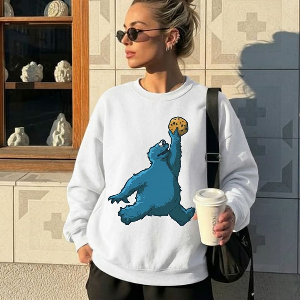 Woman wearing a white sweatshirt with a blue bear graphic, holding a coffee cup, standing in front of a decorative wall.