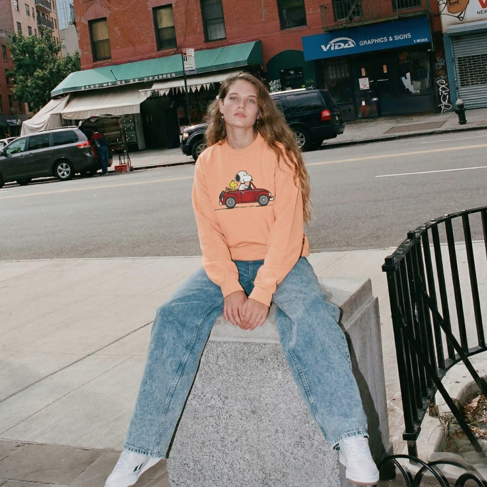 Person wearing an peach-colored sweatshirt with a car graphic sitting on a concrete block in an urban setting.
