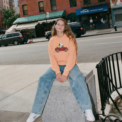 Person wearing an peach-colored sweatshirt with a car graphic sitting on a concrete block in an urban setting.