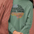 Green sweatshirt with motivational text worn by a person, against a pink background