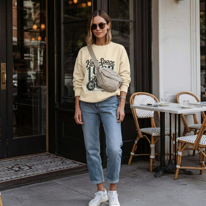 Woman in a beige sweatshirt and blue jeans standing outside a building with tables and chairs.