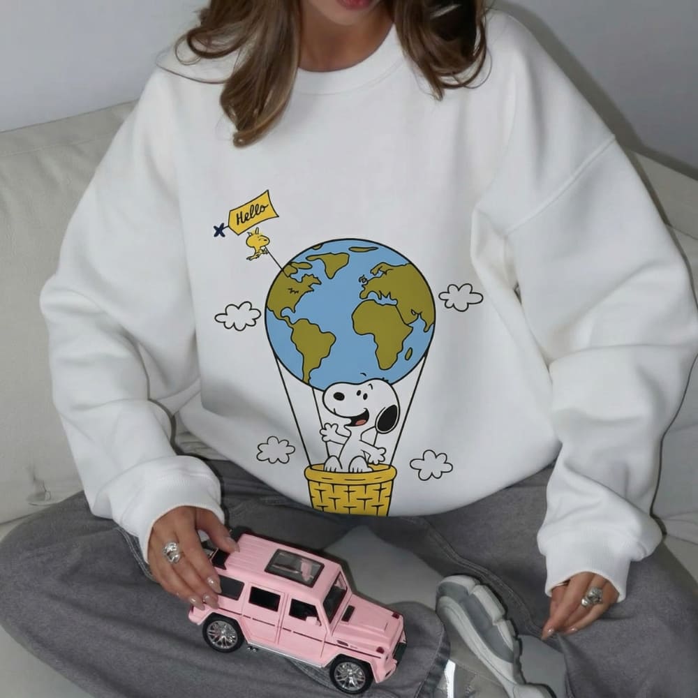 Person wearing a white sweatshirt with a cartoon design of Snoopy and the world, holding a pink toy car.
