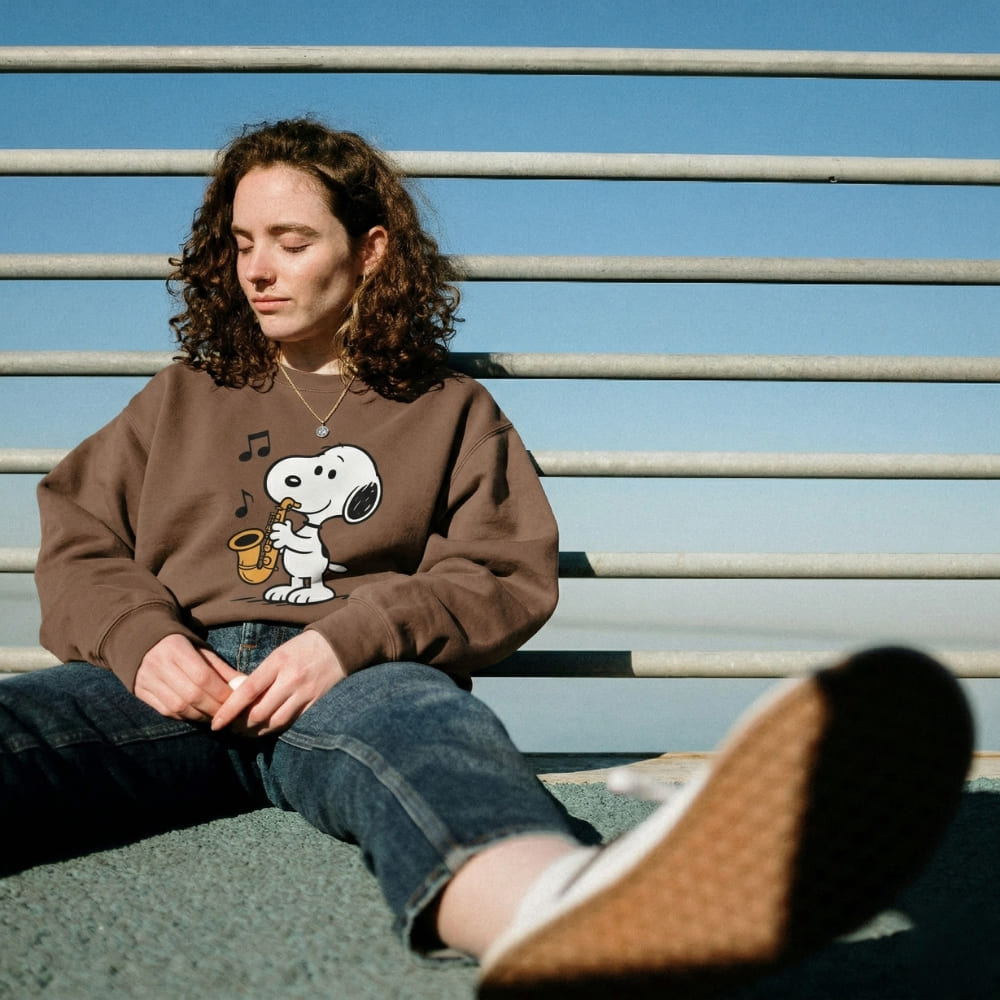 Person wearing a brown sweatshirt with a cartoon character, sitting on a bench against a blue sky.